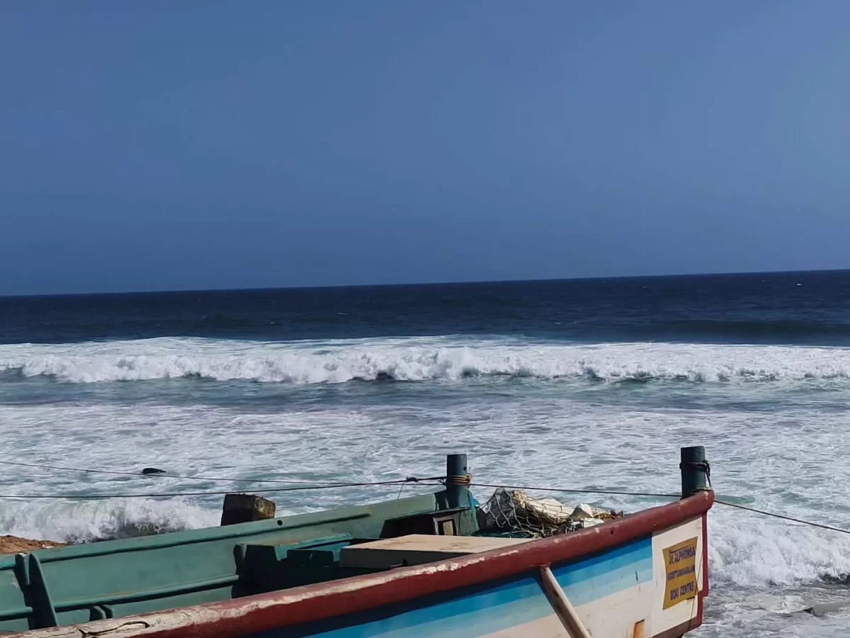 Traditional Kerala multicolored green blue red fishing boat anchored on sandy Shanghumugham Beach with crashing Arabian Sea waves under vibrant clear blue skies near Trivandrum airport, perfect South India Kerala coastal sunset tour package.