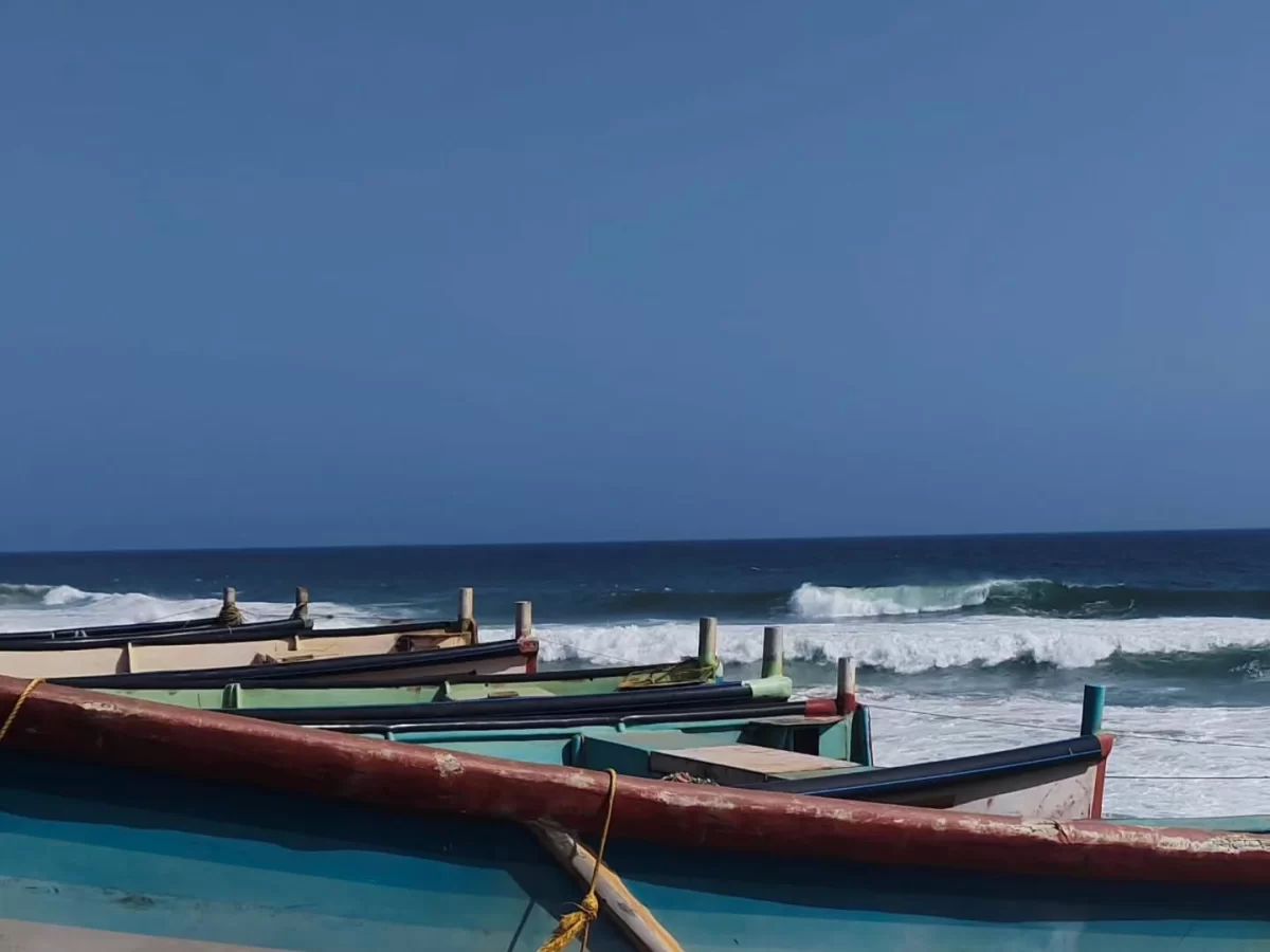 Row of colorful traditional Kerala fishing boats in vibrant green blue red hues anchored on sandy Shanghumugham Beach with Arabian Sea waves crashing under endless clear blue skies near Trivandrum, perfect South India Kerala coastal fishing village tour p