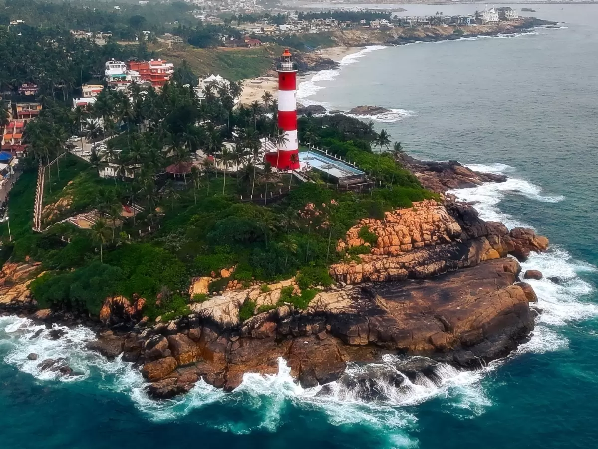 Kovalam Vizhinjam Lighthouse aerial drone view red white striped tower rocky cliff ocean waves crashing green palm trees swimming pool coastal buildings Kerala India scenic landmark tourism photography