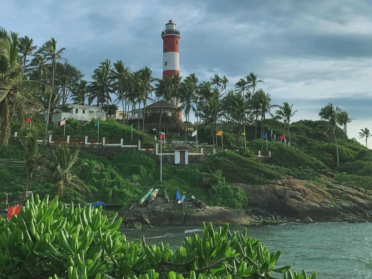 Kovalam Vizhinjam Lighthouse red white striped tower hilltop lush green vegetation palm trees flags buildings surfers ocean waves rocky coast Kerala India scenic landmark tourism photography cloudy sky.