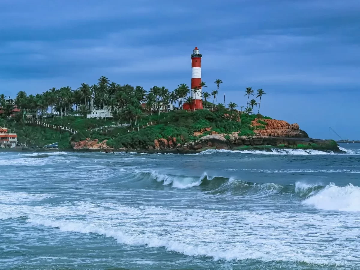 Kovalam Vizhinjam Lighthouse red white striped tower rocky green cliff palm trees ocean waves crashing under cloudy blue sky Kerala India coastal scenic landmark tourism photography dramatic weather.