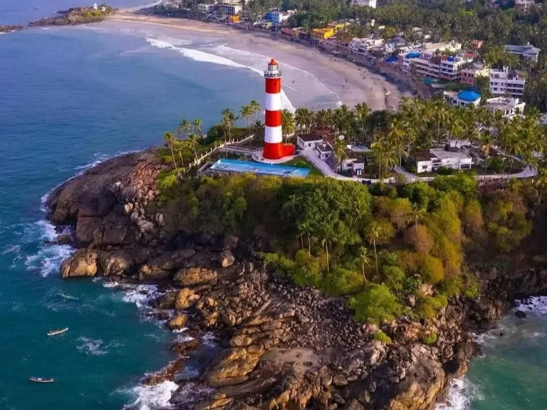 Kovalam Vizhinjam Lighthouse aerial view red white striped tower rocky green cliff swimming pool palm trees beach buildings ocean waves Kerala India coastal tourism scenic landmark drone photography.