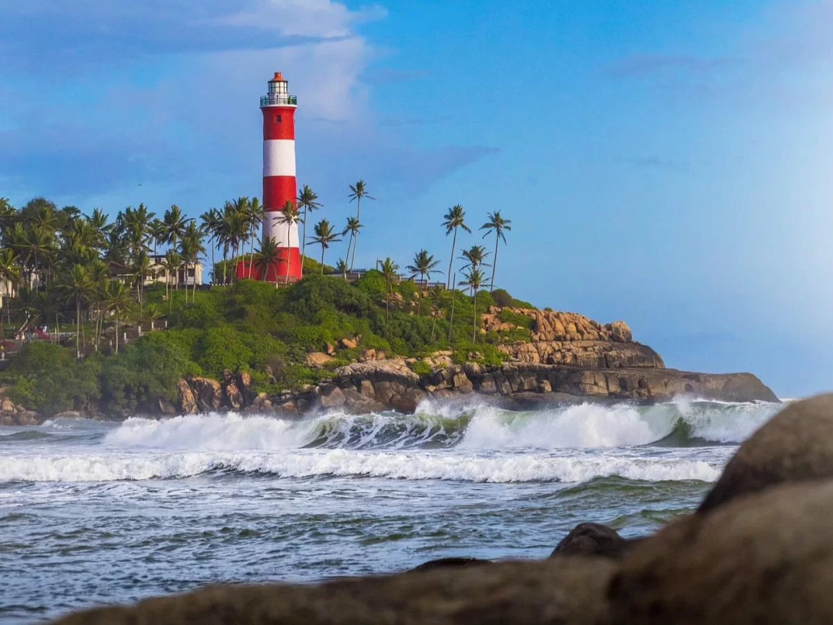 Kovalam Vizhinjam Lighthouse red white striped tower rocky green palm tree cliff ocean waves crashing foreground rocks blue sky clouds Kerala India coastal scenic tourism landmark photography golden hour light.