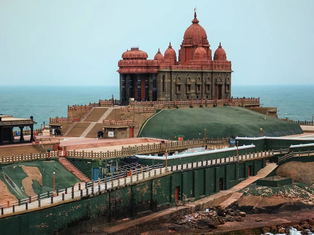 Wide angle view of Vivekananda Rock Memorial in Kanyakumari Tamil Nadu with multi-domed red shrine on green hillock, wide steps, pathways and ocean backdrop under partly cloudy sky, perfect Tamil Nadu tour package