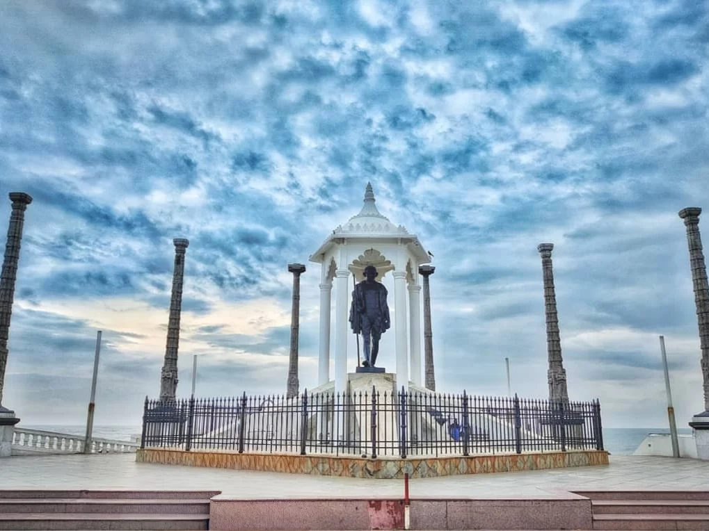 Gandhi Memorial Mandapam Kanyakumari Tamil Nadu featuring central Mahatma Gandhi statue within white pavilion columns under partly cloudy skies and beachfront plaza, essential historical patriotic Kanyakumari tour package stop