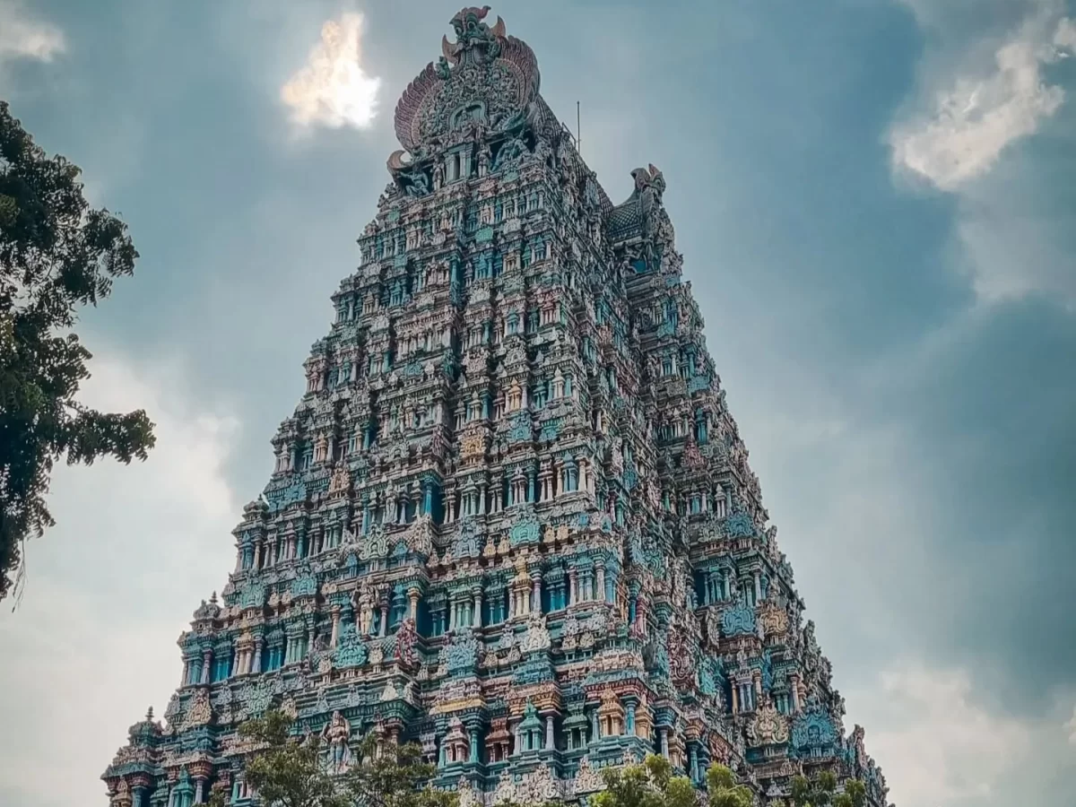 Meenakshi Amman Temple Madurai Tamil Nadu majestic soaring multi-tiered gopuram tower adorned with vibrant multicolored deity sculptures mythical motifs cloudy sky backdrop silhouetted trees, grand Dravidian Hindu temple marvel cultural heritage tour pack