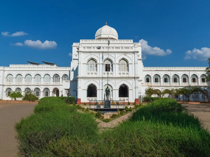 Gandhi Memorial Museum Madurai Tamil Nadu symmetrical white colonial edifice with central dome Gandhi statue foreground lush green landscaping under vibrant blue sky with clouds, freedom heritage tour package.