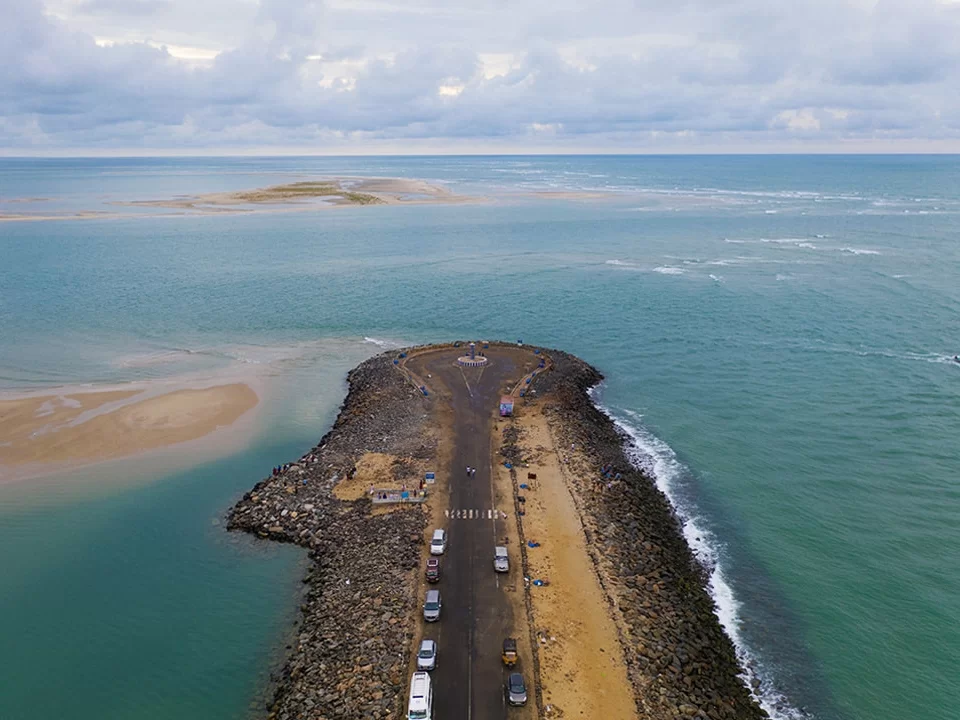 Aerial drive Pamban road at Arichal Munai Beach Dhanushkodi cloudy skies, sandbar turquoise seas distant islands vehicles jetties; perfect Rameswaram Dhanushkodi tour package. ​