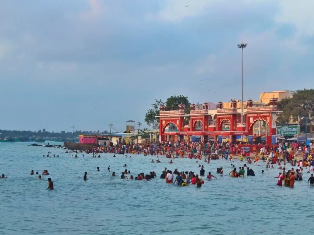 Agni Teertham Rameshwaram Ramanathapuram Tamil Nadu India vibrant red temple gate arch overlooking crowded beach pilgrims bathing sea waters cloudy skies palm trees foreground, perfect Rameswaram temple pilgrimage spiritual tour package.