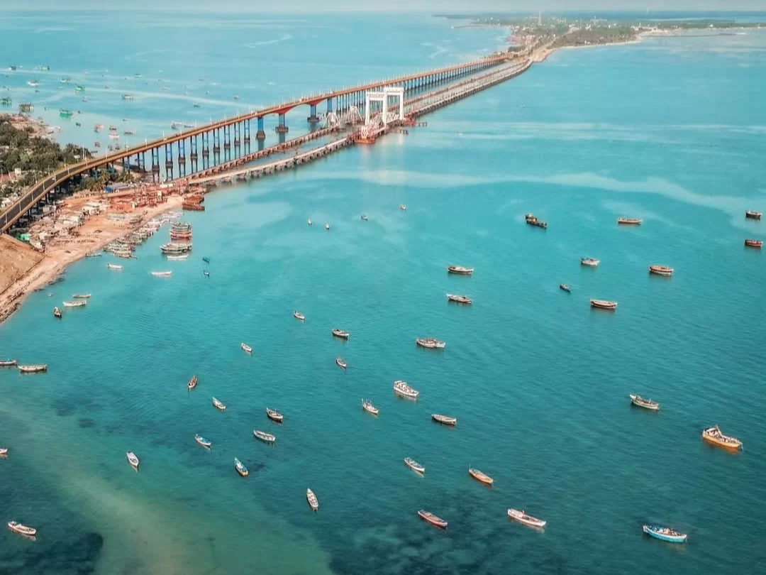 Aerial view of Pamban Bridge at Pamban Beach in Rameswaram during daytime, featuring colorful fishing boats and turquoise sea, perfect Rameswaram tour package. 