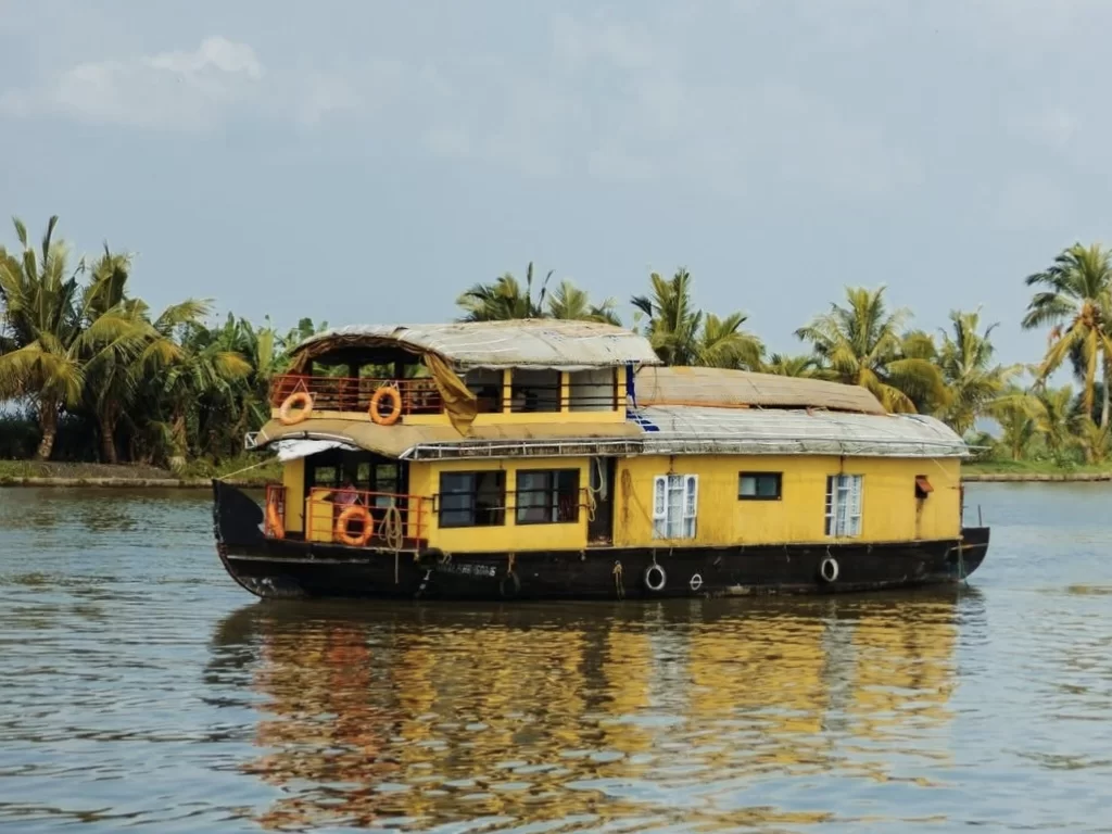 Yellow houseboat docked at Munroe Island during partly cloudy skies, featuring orange lifebuoys palms backwaters reflections, perfect romantic Kerala tour package.