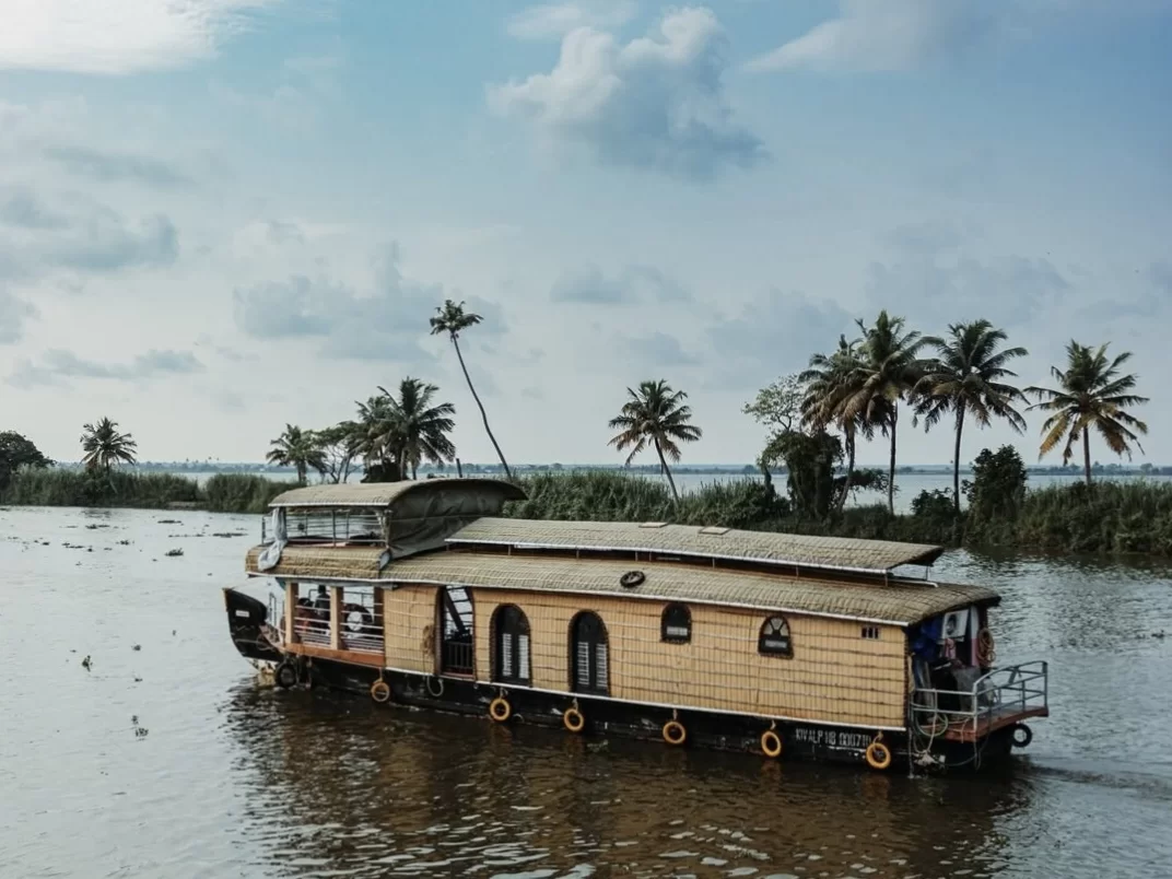 Golden houseboat sailing at Munroe Island during partly cloudy day, featuring palms backwaters passengers lifebuoys reflections, perfect romantic Kerala tour package.