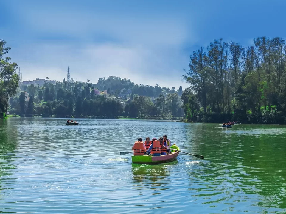 Vibrant Ooty Lake Udhagamandalam Nilgiris Tamil Nadu India boating scene with tourists in orange life jackets rowing yellow rowboat red pedal boats on turquoise waters eucalyptus shola forests distant church spire blue skies scattered clouds, perfect Nilg