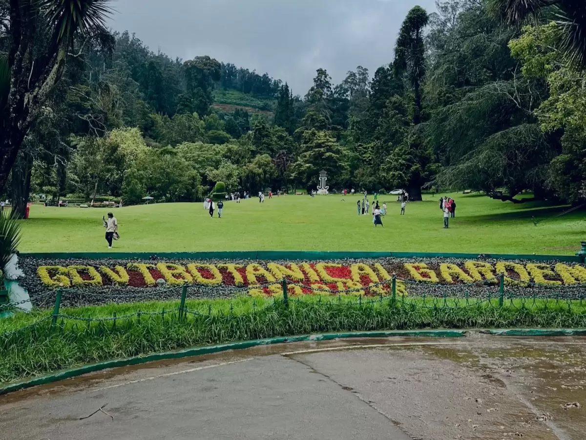 Wide view of lower garden Government Botanical Garden Ooty Udhagamandalam Nilgiris Tamil Nadu India featuring GOVT BOTANICAL GARDEN floral sign in yellow red flowers lush green Kikuyu grass lawn tourists strolling distant eucalyptus shola forest backdrop 
