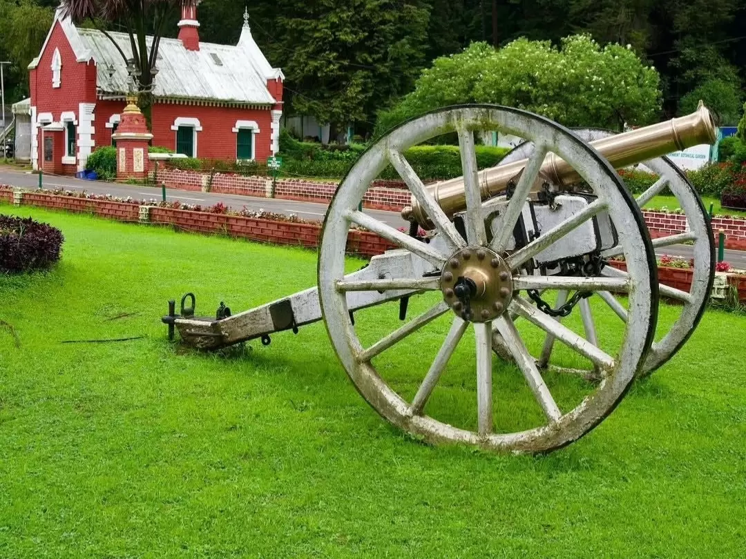 Vintage white wheel cannon on lush green lawn near red roofed white trimmed colonial gatehouse building Government Botanical Garden Ooty Udhagamandalam Nilgiris Tamil Nadu India brick boundary walls hedges misty hills backdrop, perfect Nilgiri hill statio