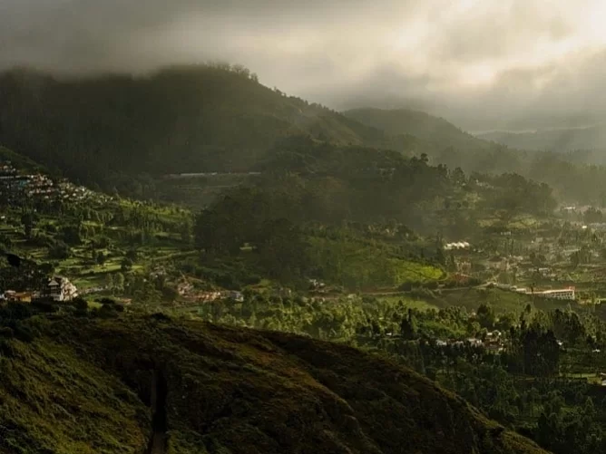 Sweeping Ketti Valley View Point Ooty Nilgiris Tamil Nadu India panorama with lush green terraced tea gardens clustered villages undulating hills shrouded in mist dramatic cloud cover golden sun rays piercing valleys, perfect Nilgiri hill station tour pac