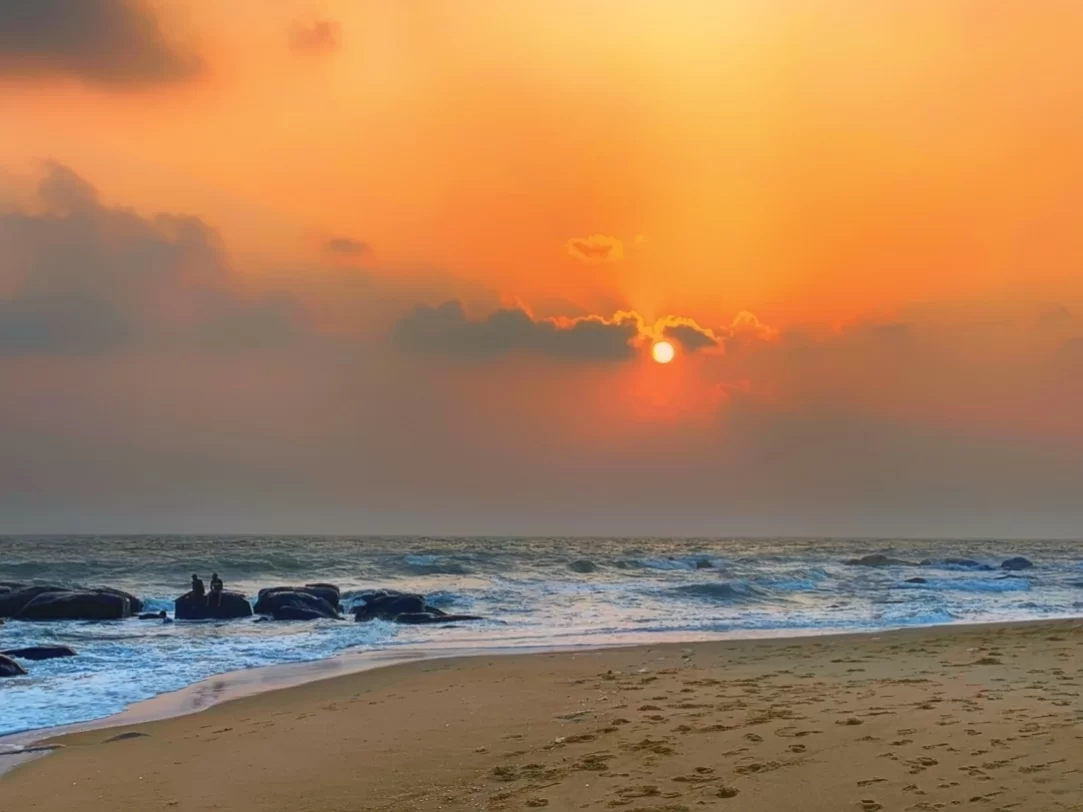Sunset at Marina Beach Chennai during golden hour, featuring orange glowing sun, crashing waves on rocks, sandy shore footprints, hazy clouds, perfect romantic experience Chennai tour package.