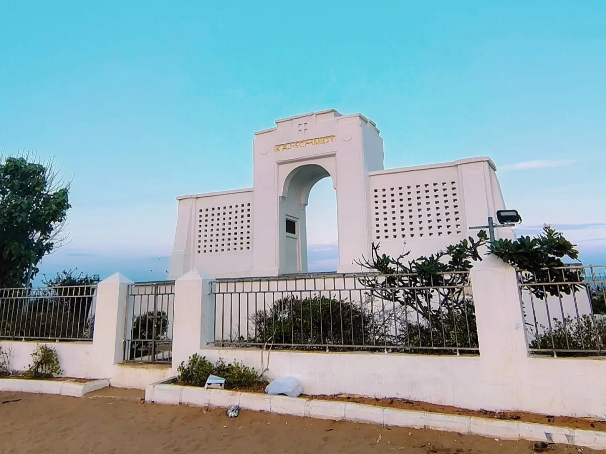 Karl Schmidt Memorial at Besant Nagar Beach during evening twilight, featuring white lattice Art Deco archway, golden Hindi inscription, palm greenery fencing, perfect coastal serenity with Chennai beach tour package.