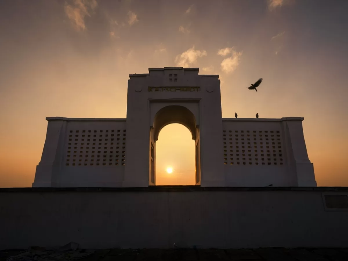 Karl Schmidt Memorial at Besant Nagar Beach during dramatic sunset silhouette, featuring white lattice archway framing sun, flying bird, cloudy skies, perfect romantic coastal experience with Chennai beach tour package.