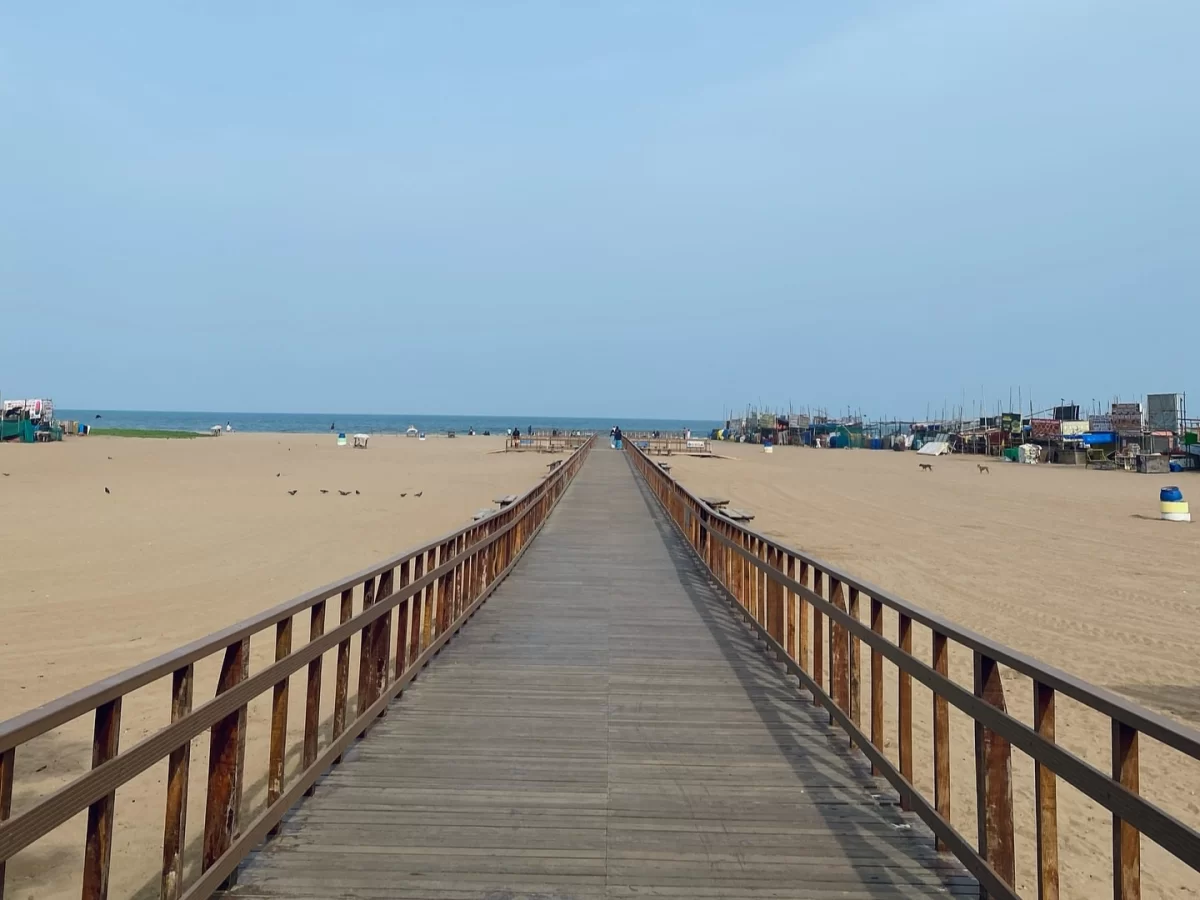 Wooden pier walk at Besant Nagar Beach during clear daytime, featuring endless golden sands, distant fishing boats, sea horizon, perfect adventure stroll with Chennai beach tour package.