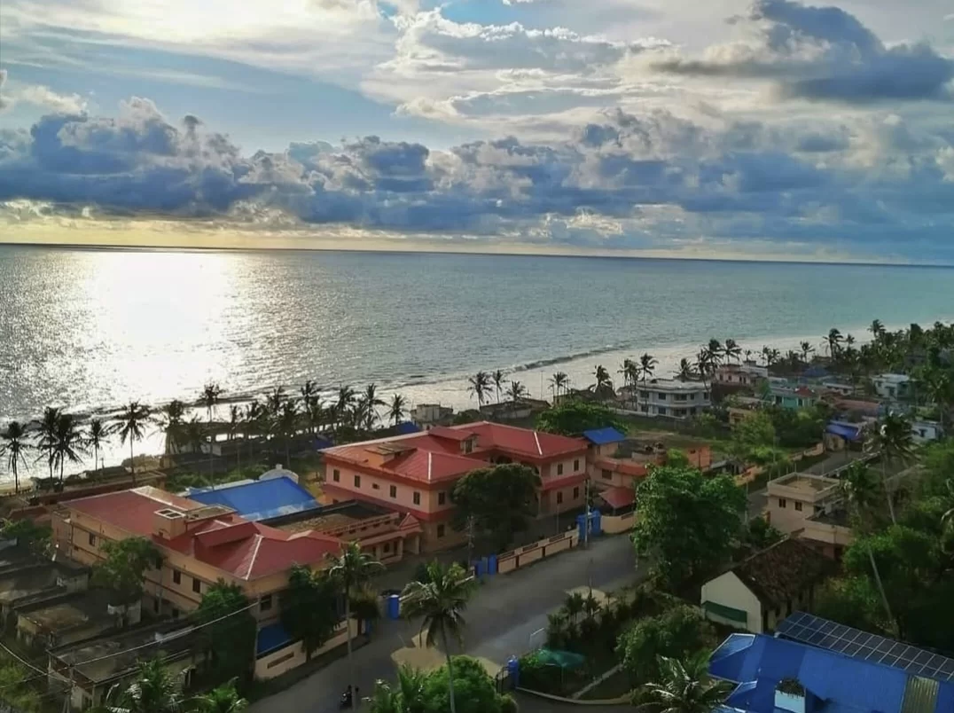 Golden hour aerial of Anjuthengu Fort Kerala village with colorful homes and resorts along pristine Arabian Sea beach under dramatic clouds, perfect romantic sunset coastal heritage stay with Varkala tour package. 
