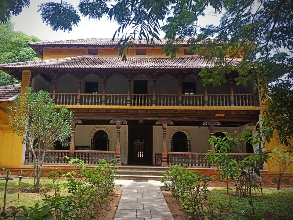 Traditional two-storey heritage house at DakshinaChitra Heritage Museum, featuring wooden balcony, tiled roof, yellow walls, and green garden pathway showcasing classic South Indian architecture.
