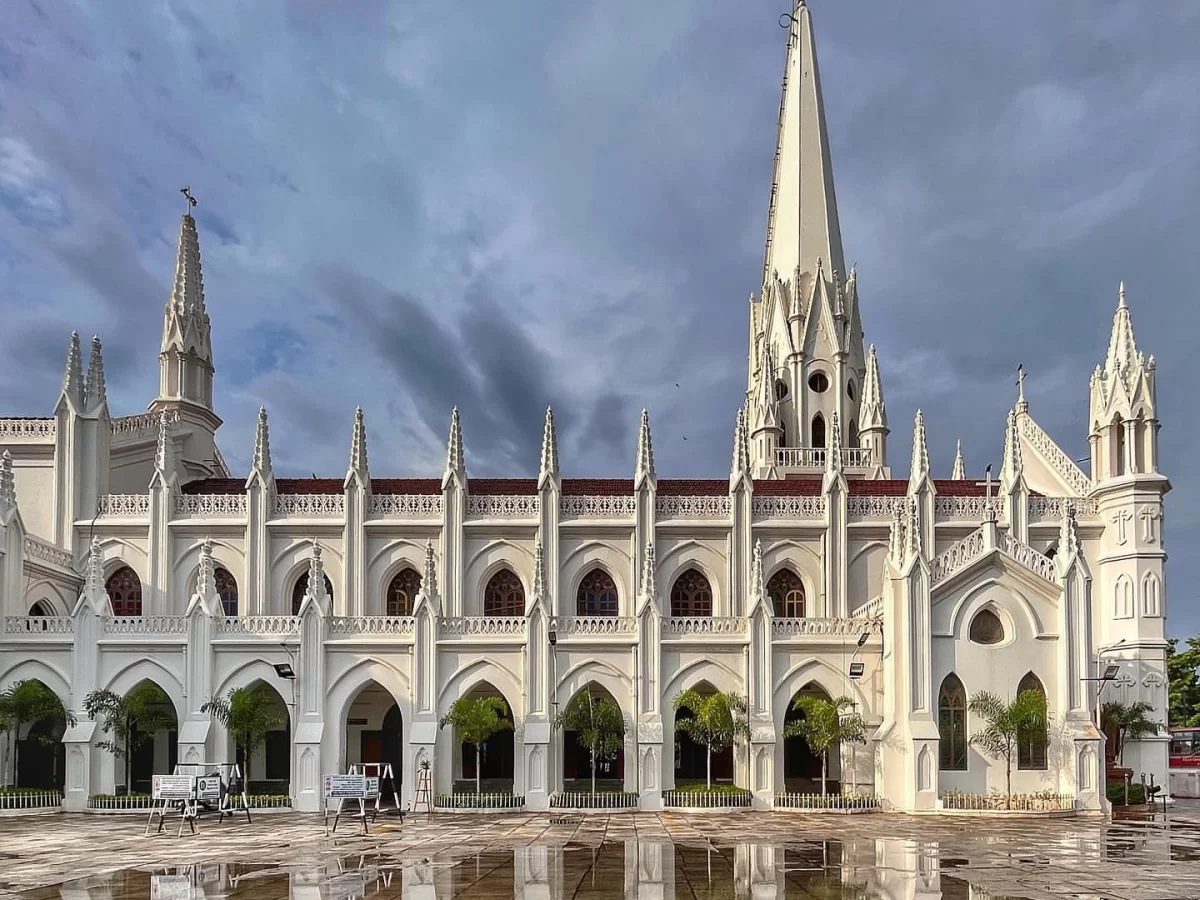 Santhome Cathedral Basilica Chennai during cloudy overcast day, featuring white Gothic arches spires, red tiled roofs, wet plaza reflections palm trees, perfect cultural experience Chennai tour package