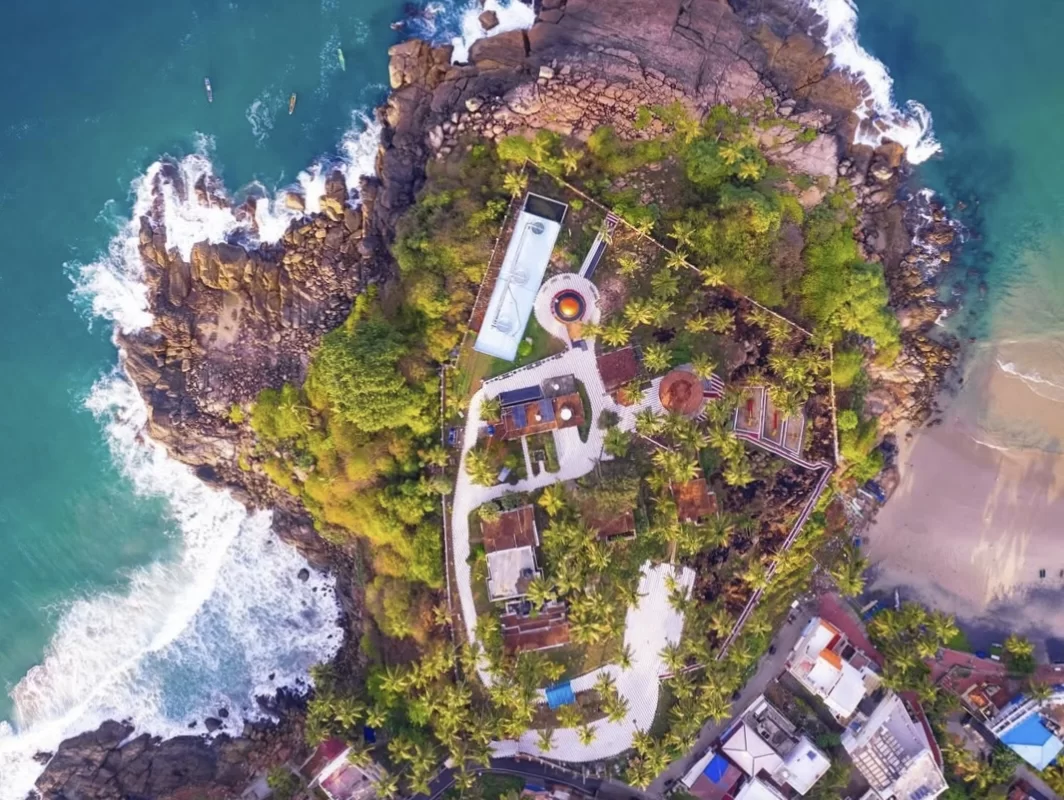 Kovalam Lighthouse Beach Kerala drone aerial view rocky green cliff ocean waves swimming pool buildings palm trees surfers luxury resort coastal scenic tourism landmark photography golden hour sunlight.