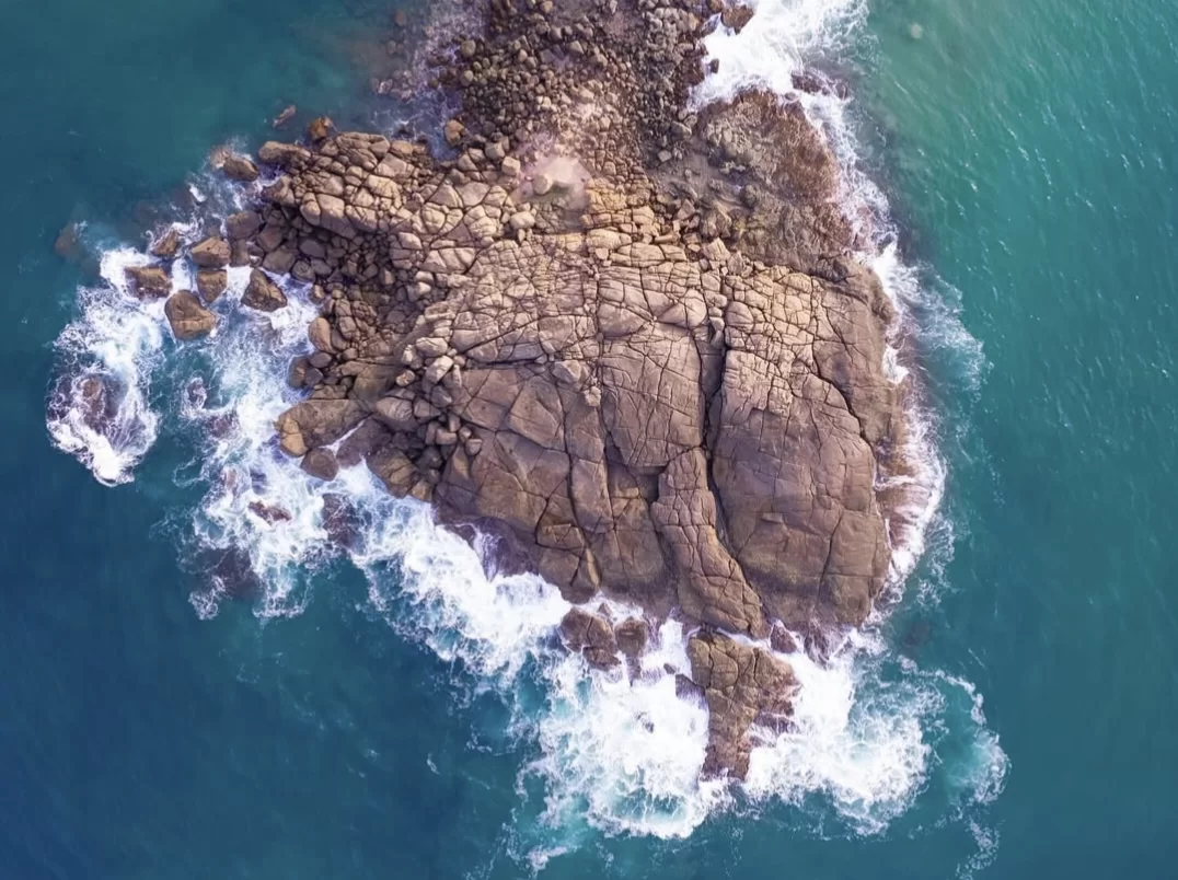 Kovalam Lighthouse Beach Kerala drone aerial view brown rocky outcrop ocean turquoise waves crashing white foam coastal scenic landmark tourism photography high tide surf dramatic sea texture.
