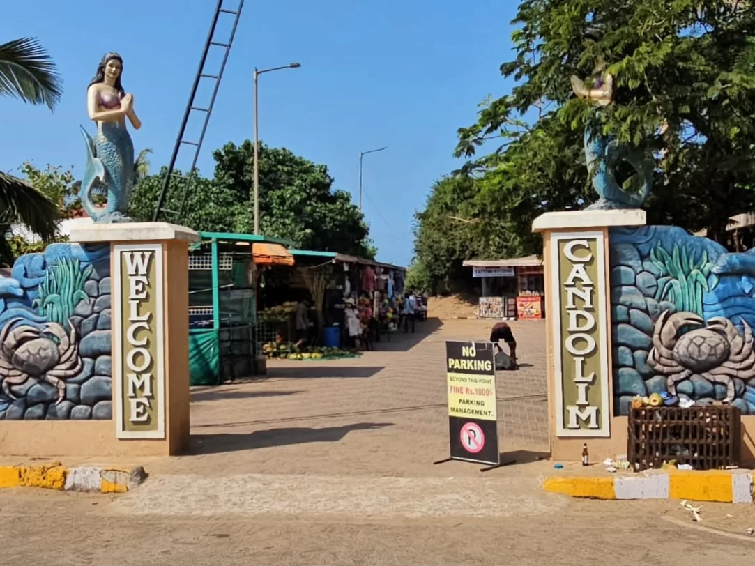 Iconic mermaid entrance gate at Candolim Beach Goa during sunny day, featuring colorful Welcome Candolim signage crabs parking sign shops trees, perfect beach access experience with Candolim Beach Goa tour package. 