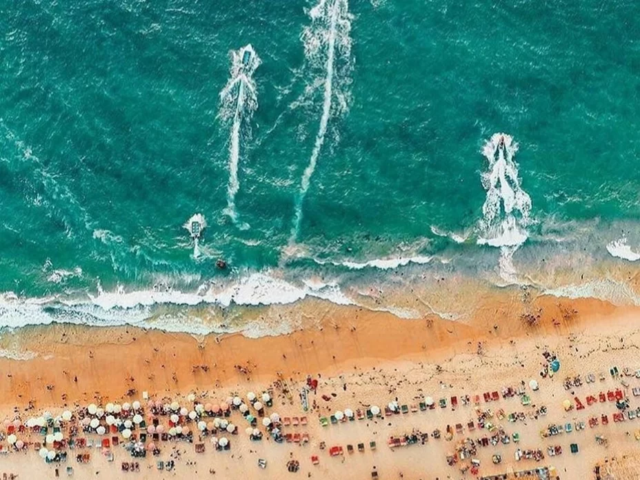 Aerial view of jet skis trails on turquoise Arabian Sea at Candolim Beach Goa during sunny day, featuring crowded sandy shore colorful umbrellas chairs waves, perfect water adventure experience with Candolim Beach Goa tour package.