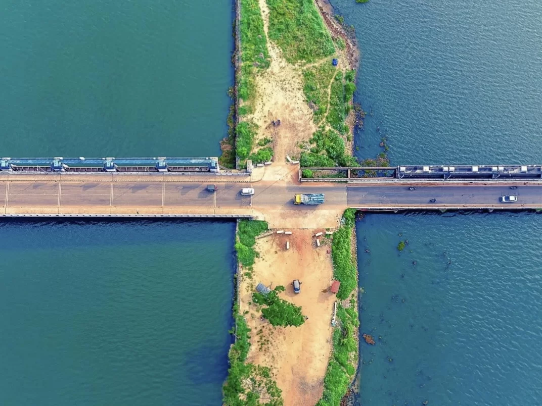 Drone aerial of Thannermukkom Bund across Vembanad Lake Kerala during calm day, featuring road bridge, vehicles, V-jetties, lush banks, turquoise waters, perfect backwater experience with Alleppey tour package.