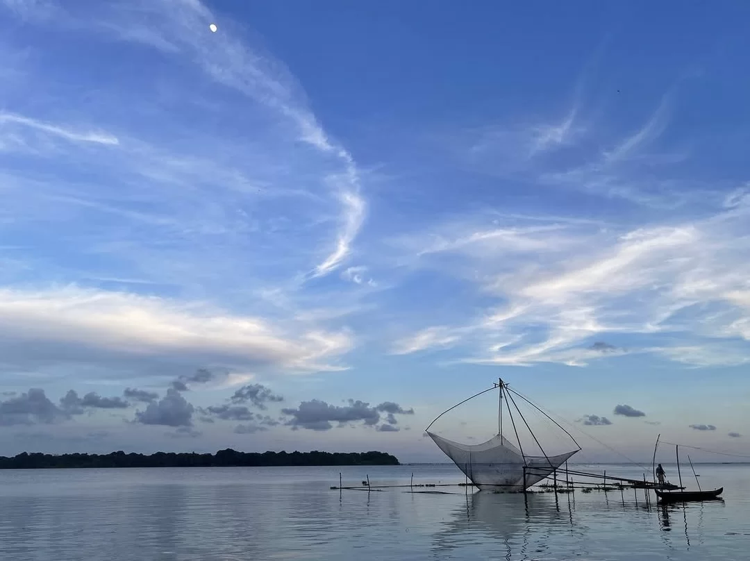 Chinese Fishing Net on Vembanad Lake Kerala at twilight, featuring iconic white cheena vala net, crescent moon, wispy clouds, serene waters, distant greenery, boats, perfect cultural backwater experience with Alleppey houseboat tour package.