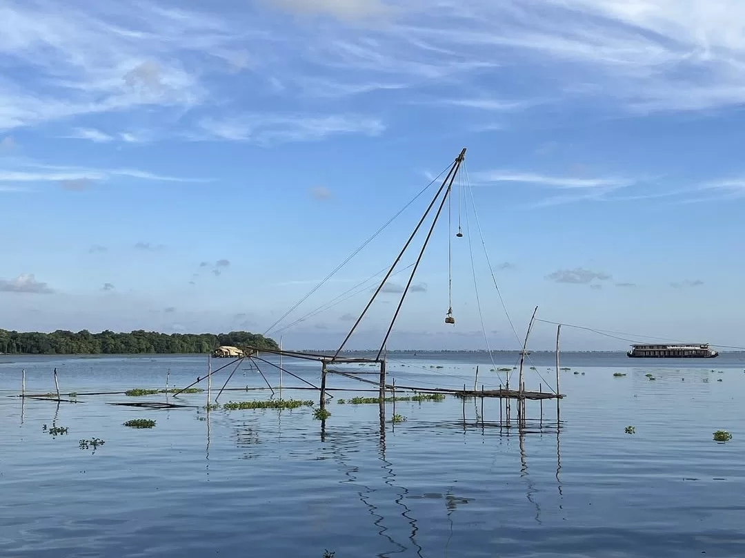 Traditional Chinese Fishing Net on Vembanad Lake Kerala during daytime, featuring wooden cantilever structure with pulley, calm backwaters, water hyacinth, distant palms, houseboat, perfect authentic fishing heritage experience with Alleppey backwater tou