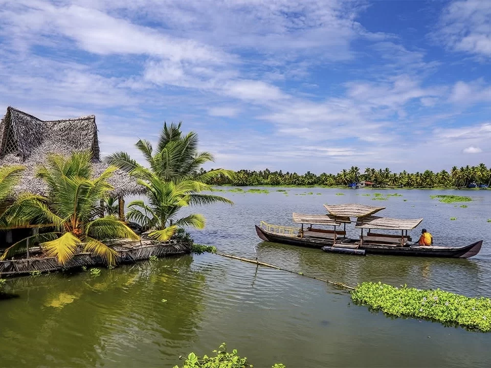 Traditional thatched homestay and kanawa boat on Vembanad Lake backwaters Kerala under partly cloudy sky, featuring palm trees, water hyacinth, lush greenery, perfect authentic rural experience with Alleppey village backwater tour package. 