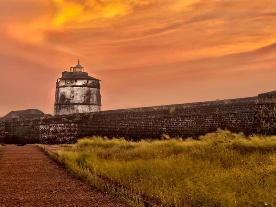 Sunset view of Fort Aguada lighthouse tower bastion walls Candolim Goa during vibrant orange sky, featuring weathered laterite structure golden grass pathway, perfect romantic heritage experience with Fort Aguada Goa tour package.