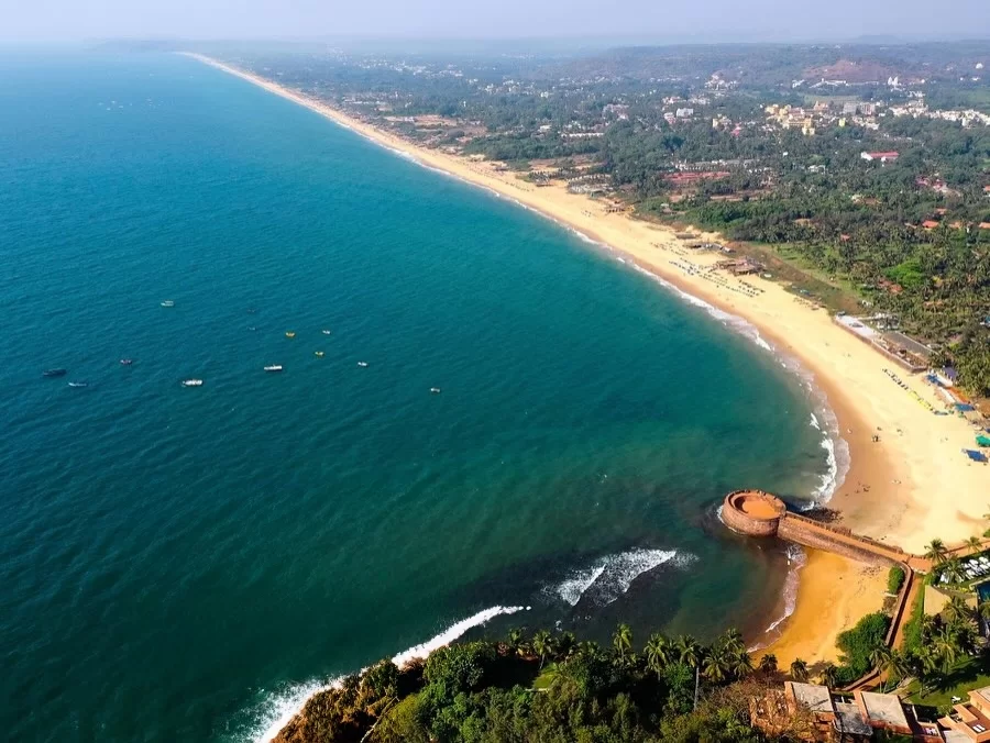 Aerial view of Sinquerim Beach in Goa during sunny day, featuring Aguada Fort bastion, golden sands, Arabian Sea waves and palm groves, perfect beach experience with Goa tour package. 
