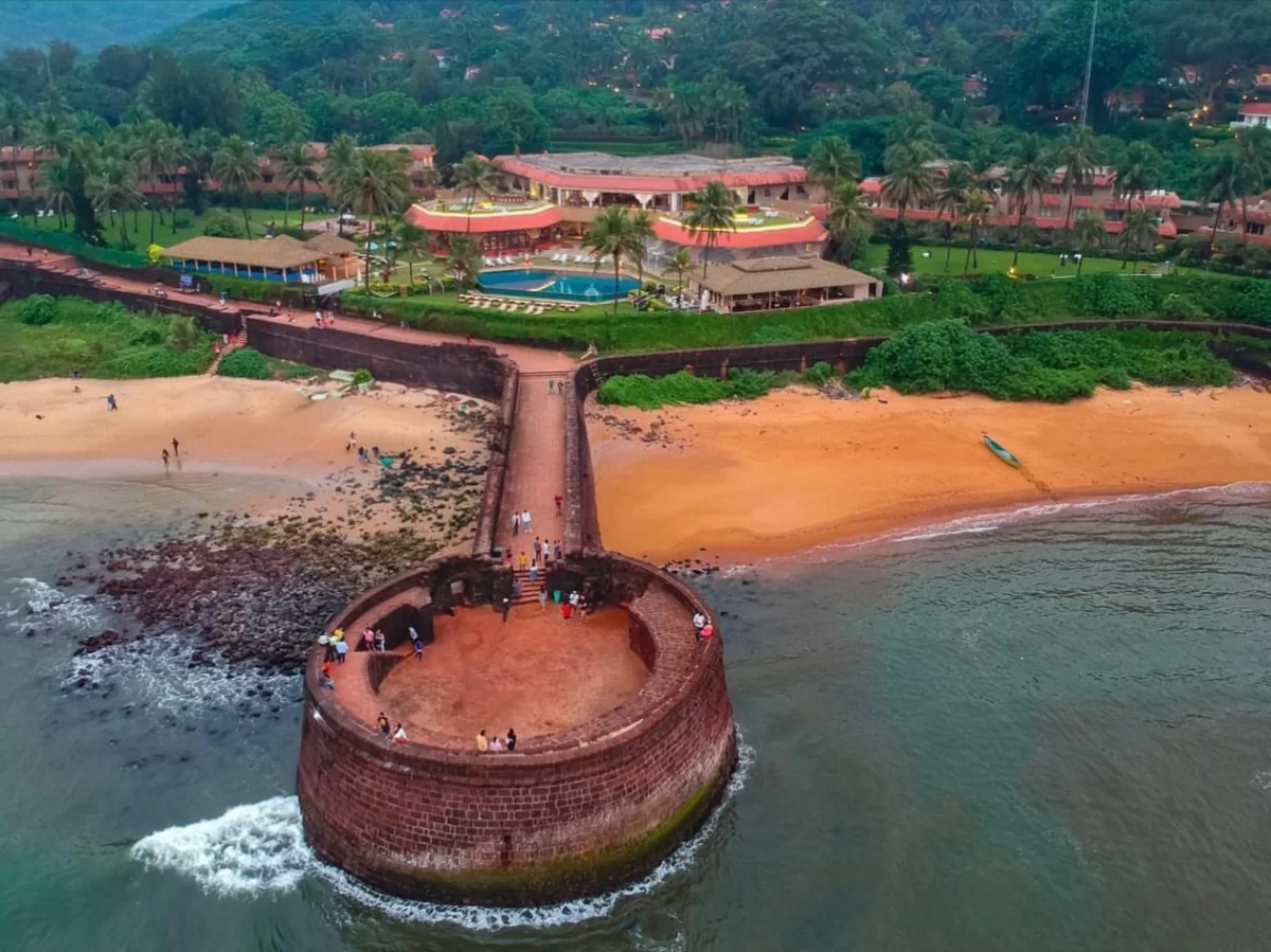 Drone aerial view of Fort Aguada complex Sinquerim Beach Candolim Goa during monsoon, featuring red laterite bastion jetty swimming pool luxury resort palm trees Arabian Sea waves sandy shores tourists greenery, perfect panoramic luxury heritage experienc