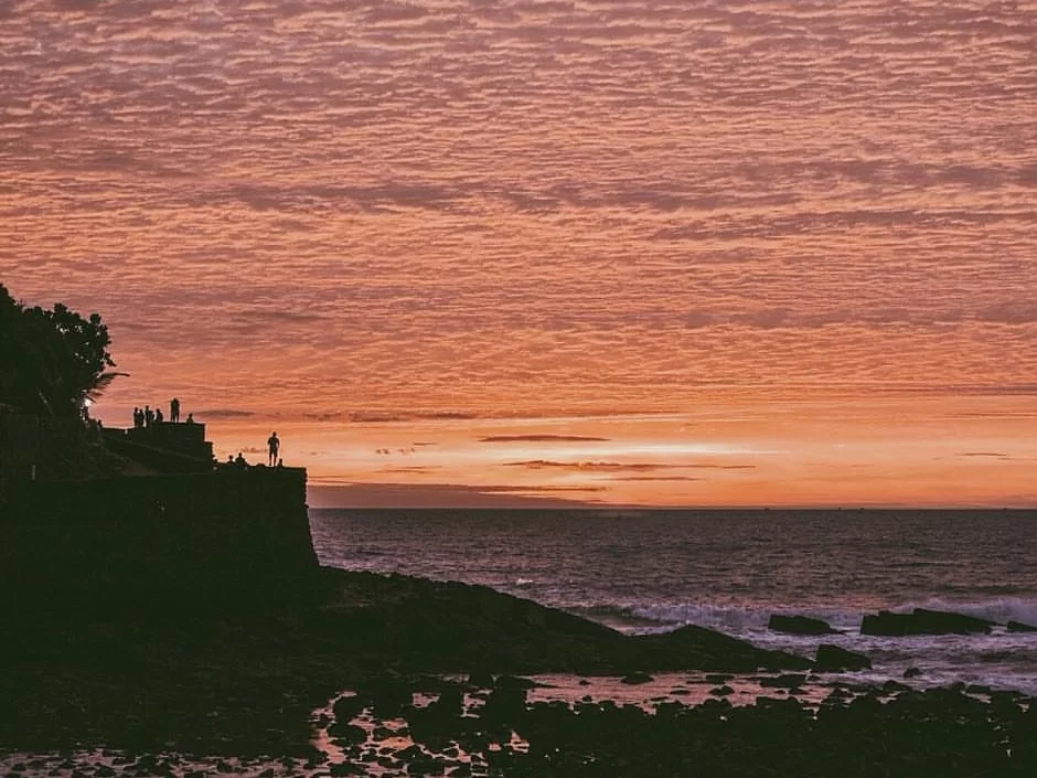 Silhouettes on cliff at Sinquerim Beach Goa during vibrant sunset, featuring crashing waves and orange skies, perfect romantic experience with Goa tour package. 