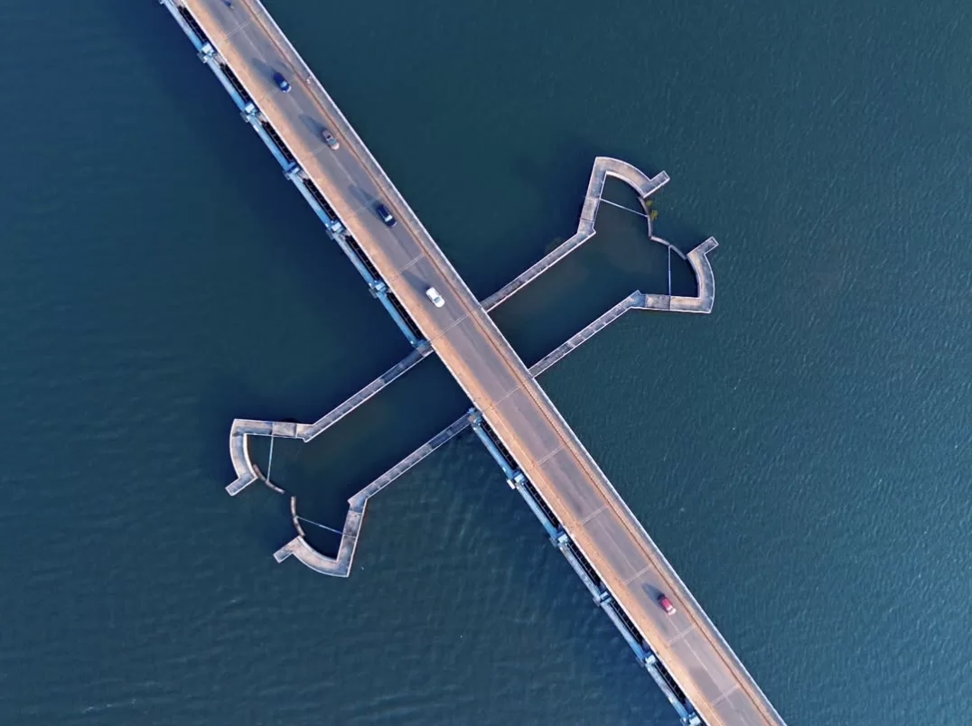 Aerial view of Thannermukkom Bund Vembanad Lake during clear day, featuring cross-shaped barrage, V-jetties, vehicles, deep blue waters, perfect backwater experience with Alleppey tour package