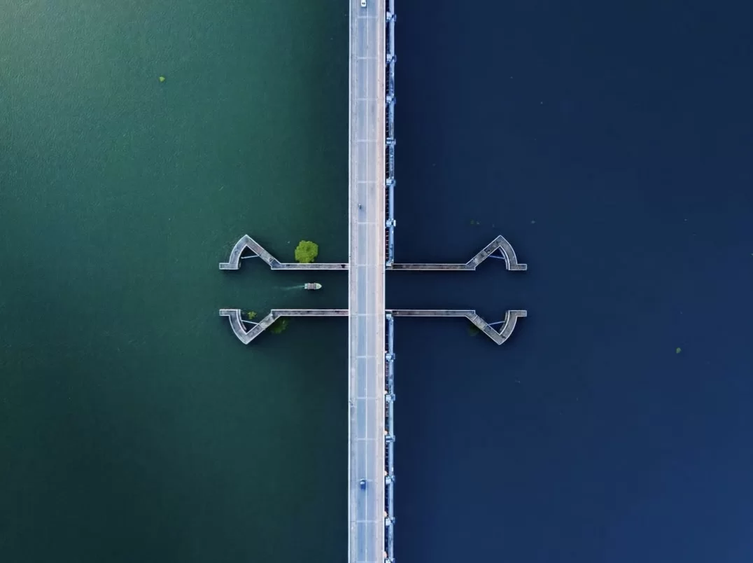 Aerial drone view of Thannermukkom Bund Vembanad Lake during sunny day, featuring saltwater barrier dividing green-fresh waters, V-jetties, road bridge, perfect backwaters experience with Kumarakom tour package. 