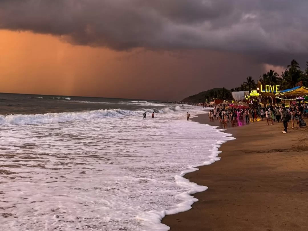 Goa Anjuna Beach golden hour dramatic orange purple stormy clouds yellow LOVE beach shack sign crowd tourists colorful sarees people walking swimming sandy shore ocean waves palm trees coastal party vibe sunset
