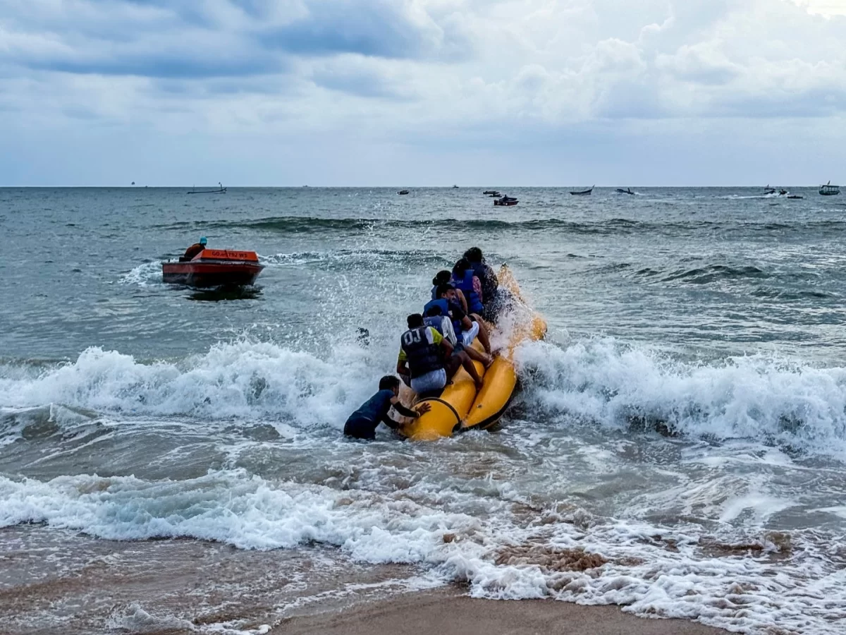 Goa Anjuna Beach banana boat ride yellow inflatable boat group blue shirt tourists splashing waves speedboat pulling orange life rings distant boats cloudy sky sandy beach water adventure sports tourism fun activity.