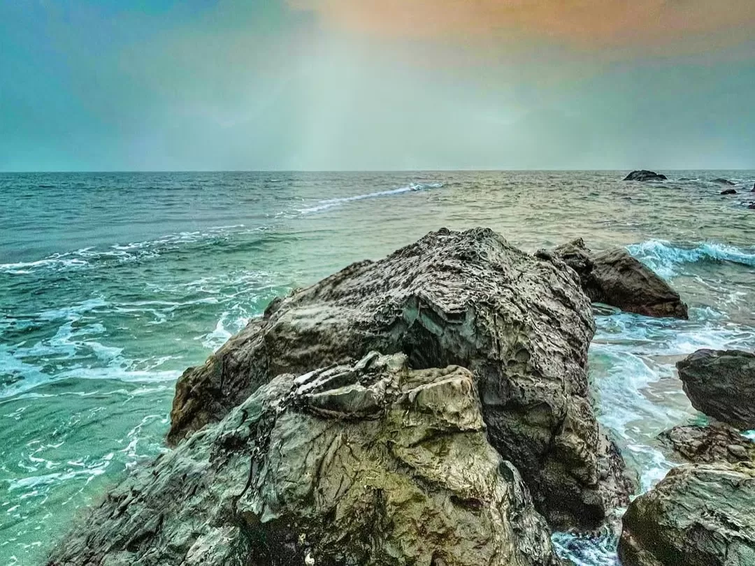 Dramatic Vagator Beach Goa seascape with turquoise waves crashing on massive rocky outcrops under split blue-orange sunset sky, perfect adventurous cliffside beach vibe with North Goa tour package.