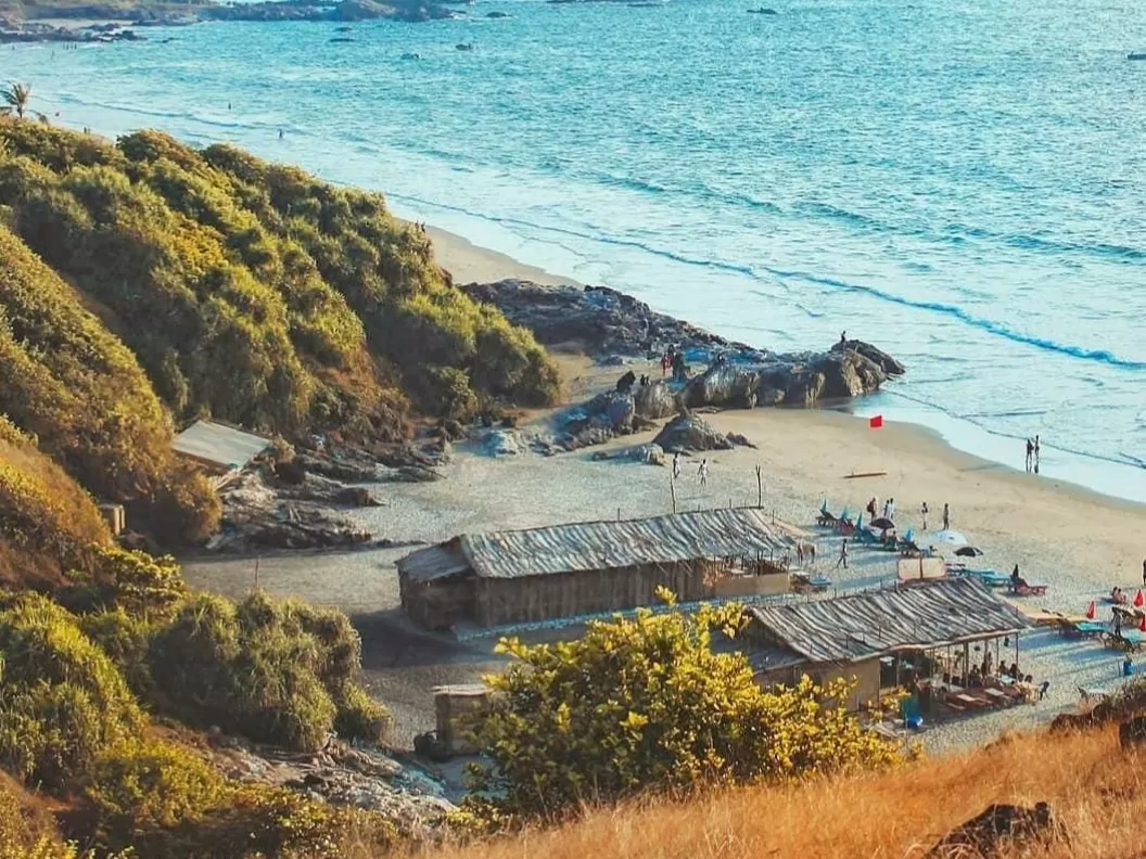 Vibrant Vagator Beach Goa from cliffside with shack beach huts, umbrellas, rocky shoreline and azure Arabian Sea under golden light, perfect boho beach shacks vibe with North Goa tour package. 