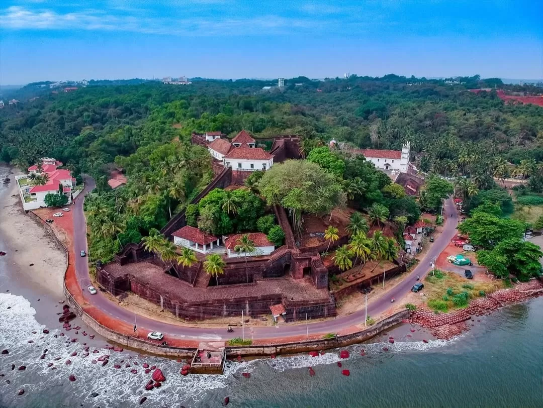 Aerial view of Reis Magos Fort in Goa during clear day, featuring red laterite walls, white churches, lush greenery and Arabian Sea coastline, perfect heritage experience with Goa tour package. 