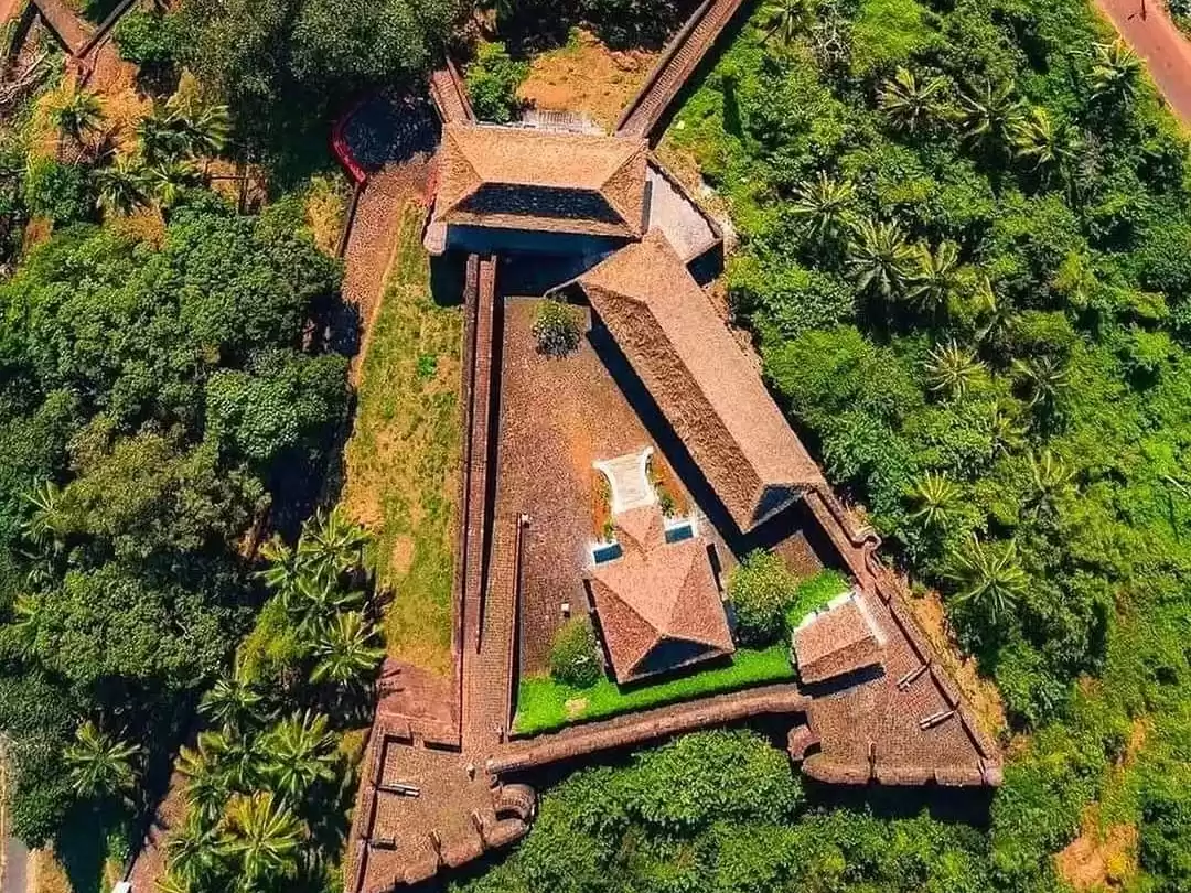 Aerial view of Chapora Fort in Goa during sunny day, featuring red laterite ramparts, terracotta roofs, lush greenery and palm trees, perfect adventure experience with Goa tour package. 
