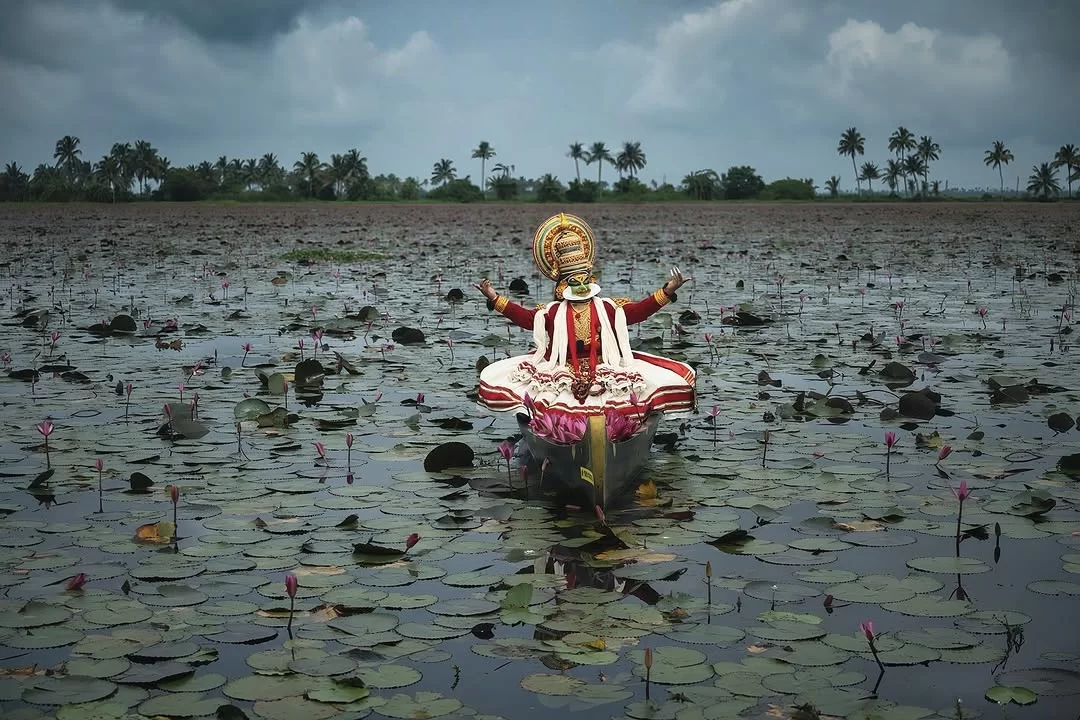 Kerala Vembanad Lake Kathakali artist golden crown green face red white elaborate costume arms outstretched black boat pink lotus blooms green lily pads coconut palms cloudy sky, cultural performance backwaters tour package