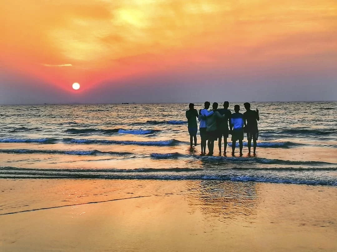 Group of silhouetted friends watching golden sunset over Arabian Sea at Miramar Beach Goa, featuring orange sky gentle waves sandy shore, perfect beach experience Goa sunset beach outing package.