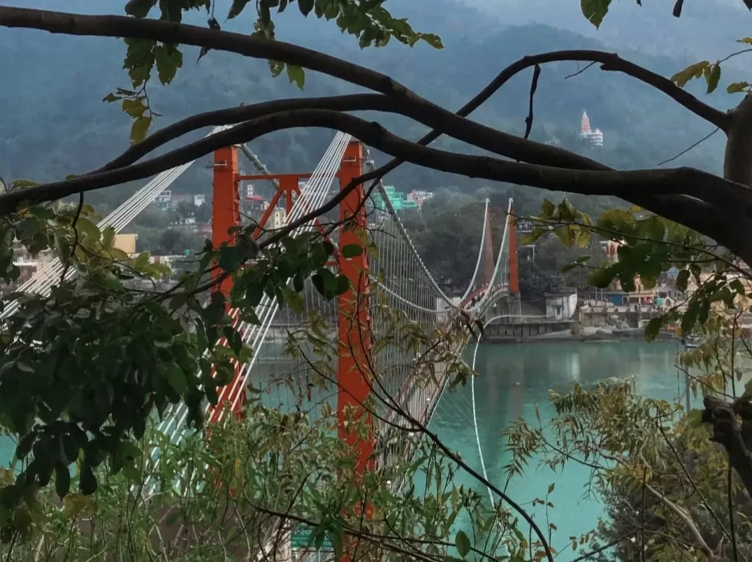 Laxman Jhula new suspension bridge at Rishikesh over Ganga River, through trees featuring red towers cables Neelkanth Temple hilltop buildings, perfect spiritual experience Uttarakhand tour package.